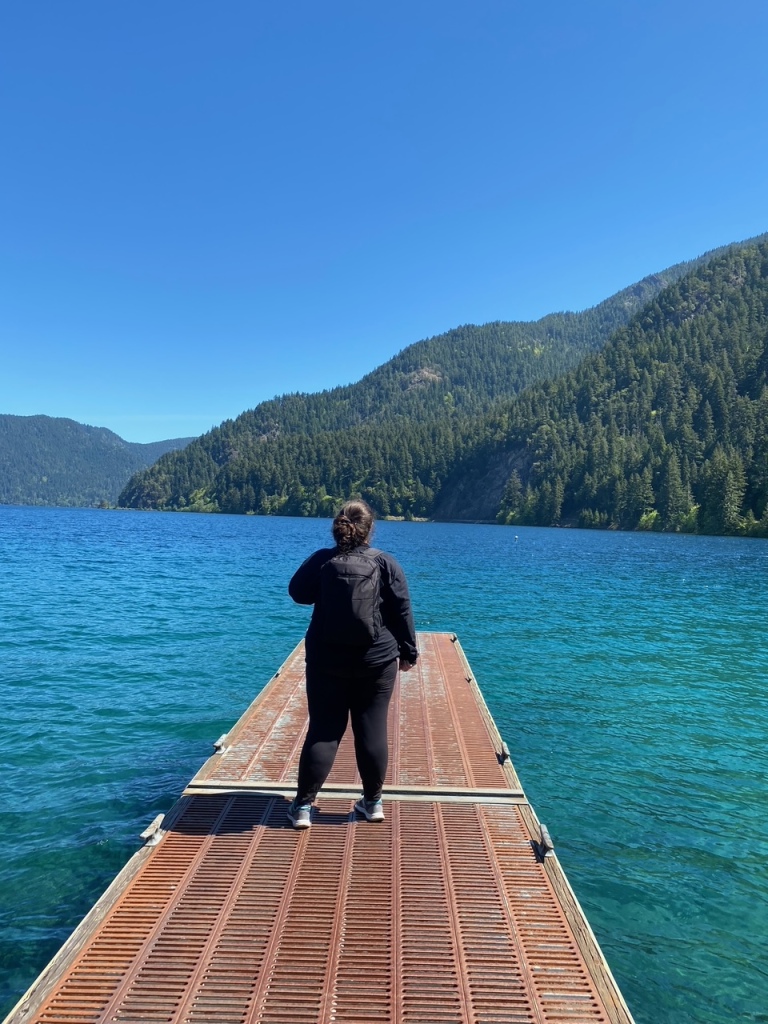 A chubby woman in black with a black backpack stands on an oragne dock and faces away from the camera, looking out at bright blue-green water and green mountains, topped with a deep blue sky.